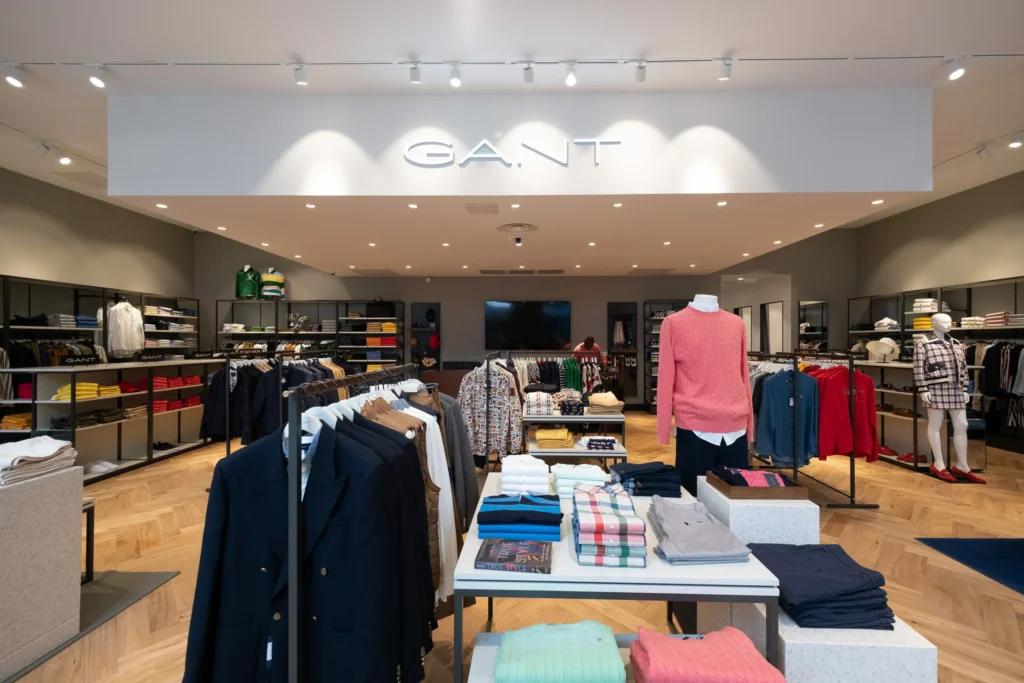 Vue de face d’une boutique GANT avec logo lumineux, tables produits, vêtements et rayons muraux