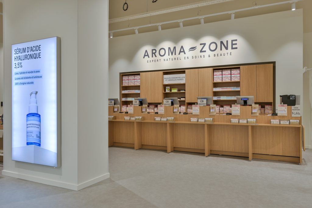 Espace caisse Aroma-Zone Anglet avec logo mural, comptoir bois et affichage digital produit