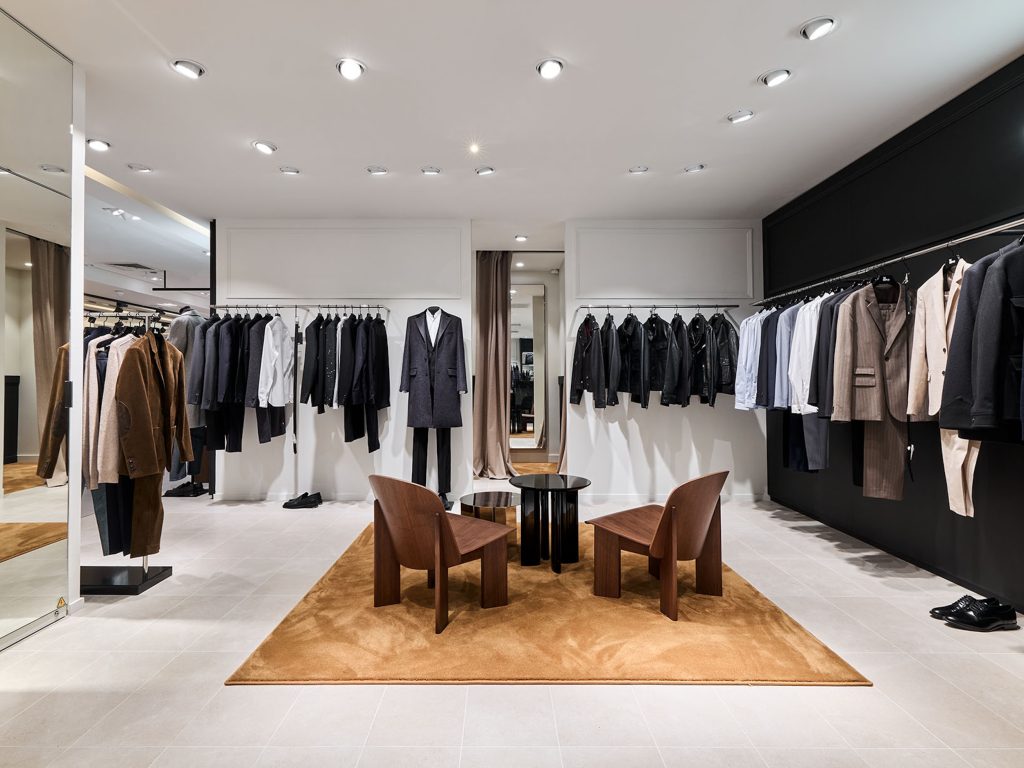 Espace essayage du corner Homme The Kooples avec fauteuils bois, tapis brun, portants et vêtements premium