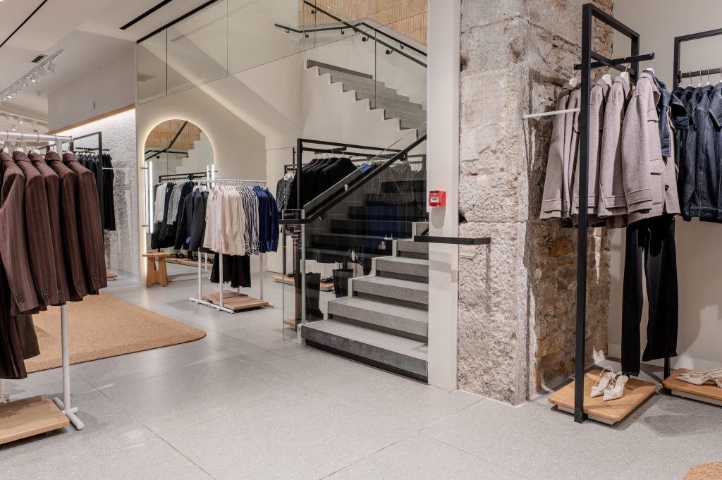 Escalier intérieur de la boutique MANGO Lyon République avec garde-corps vitré, murs en pierre et portants textile