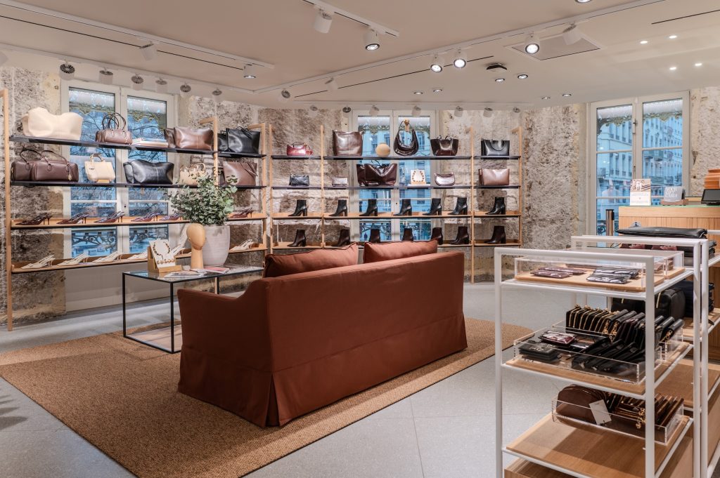 Linéaires de sacs et chaussures dans la boutique MANGO Lyon avec murs en pierre, canapé et mobilier bois