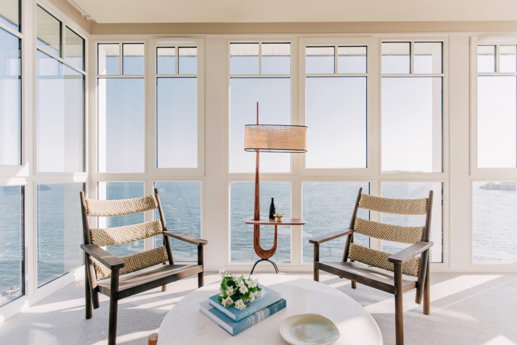 Salon lumineux de l’hôtel Les Bassans avec fauteuils, lampadaire et grandes baies vitrées face à la mer
